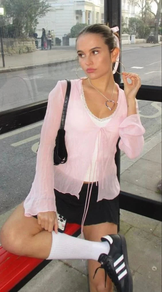 Young woman in a coquette-inspired outfit, wearing a sheer pink blouse, black mini skirt, knee-high socks, and Adidas sneakers, sitting at a bus stop in an urban setting.