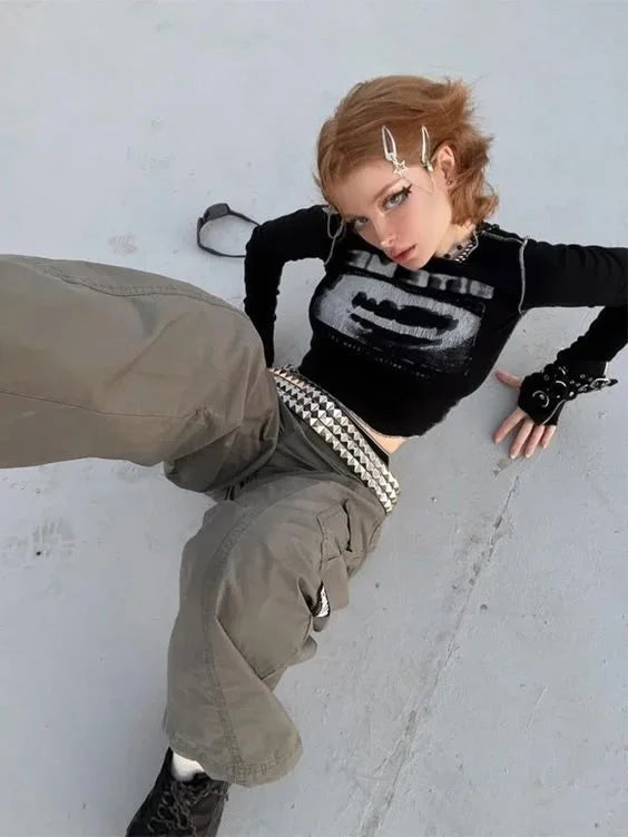 Young woman in a fairy grunge-inspired outfit, wearing a distressed black crop top, oversized cargo pants, layered chains, studded belt, and statement hair clips, posing on a concrete surface with an ethereal yet edgy vibe.