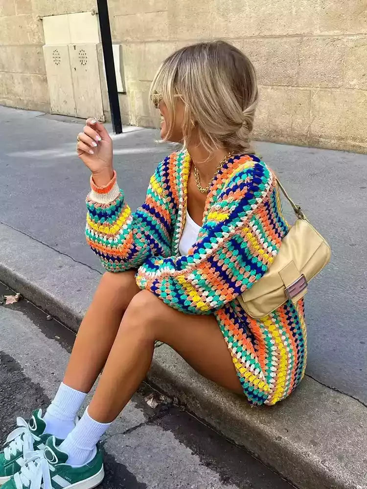 
Young woman in a colorful, retro-inspired outfit, wearing a vibrant crochet cardigan, white tank top, mini skirt, green sneakers, and a cream-colored shoulder bag, sitting casually on the sidewalk with a relaxed, effortless vibe.